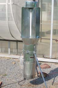 A black light trap, with a shiny metal exterior, stands guard outside a greenhouse.